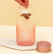 Aufgeschraubten Ice Tea Flasche in Rosa, in der eine Hand einen Tree of Tea Teebeutel hineinführt.