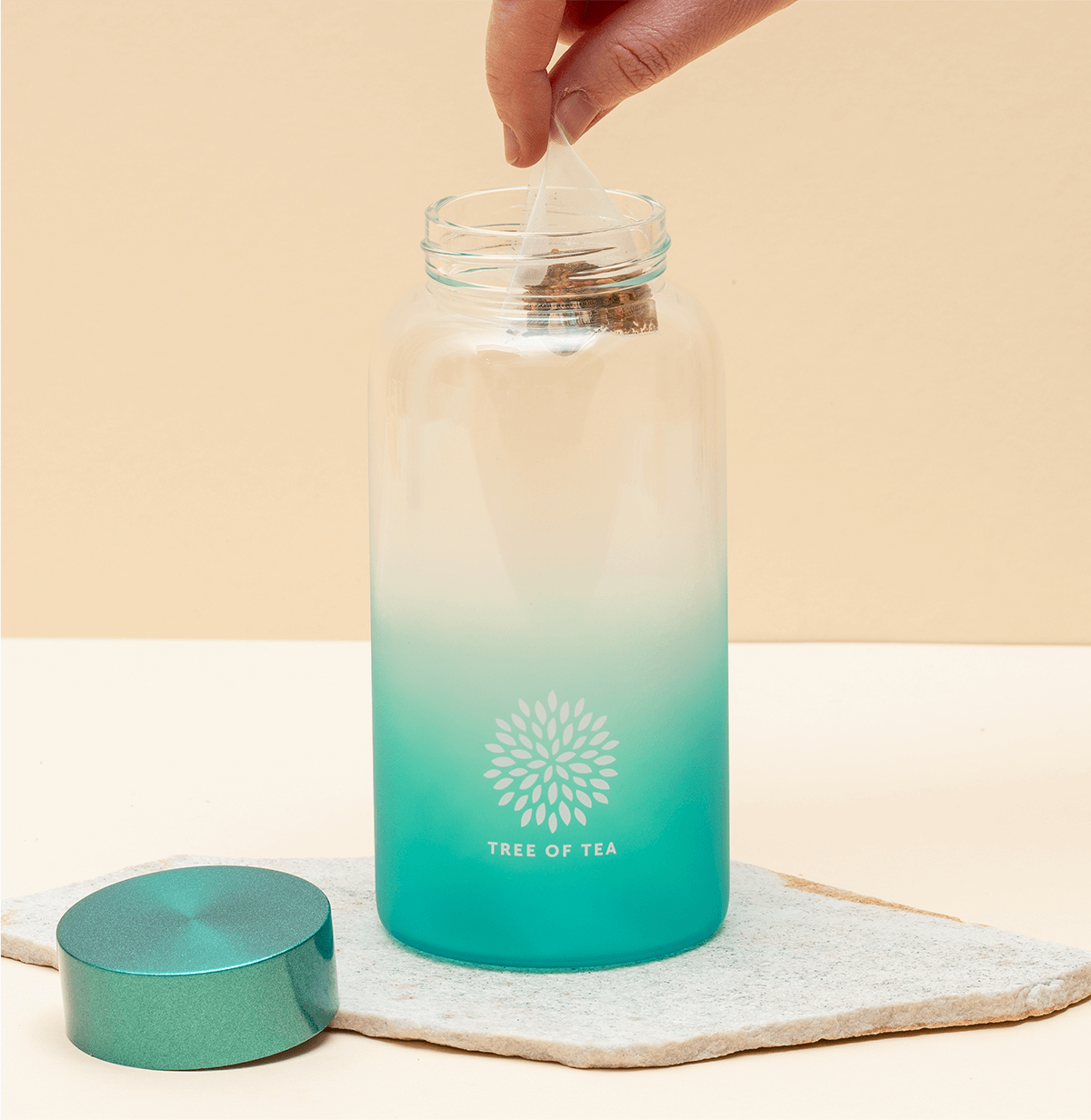Aufgeschraubten Ice Tea Flasche in Türkis, in der eine Hand einen Tree of Tea Teebeutel hineinführt.