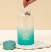 Aufgeschraubten Ice Tea Flasche in Türkis, in der eine Hand einen Tree of Tea Teebeutel hineinführt.