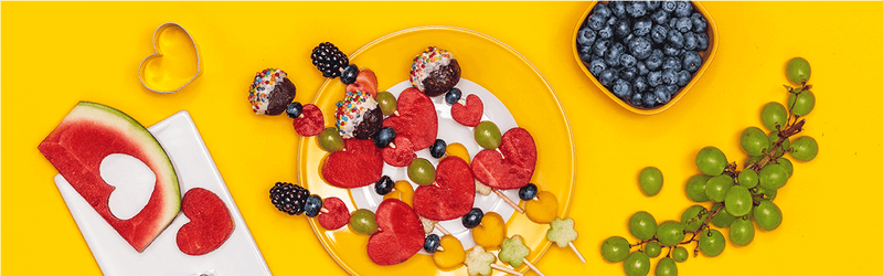Fruchtspieße für Kinder auf einem Teller mit Weintrauben und Blaubeeren daneben