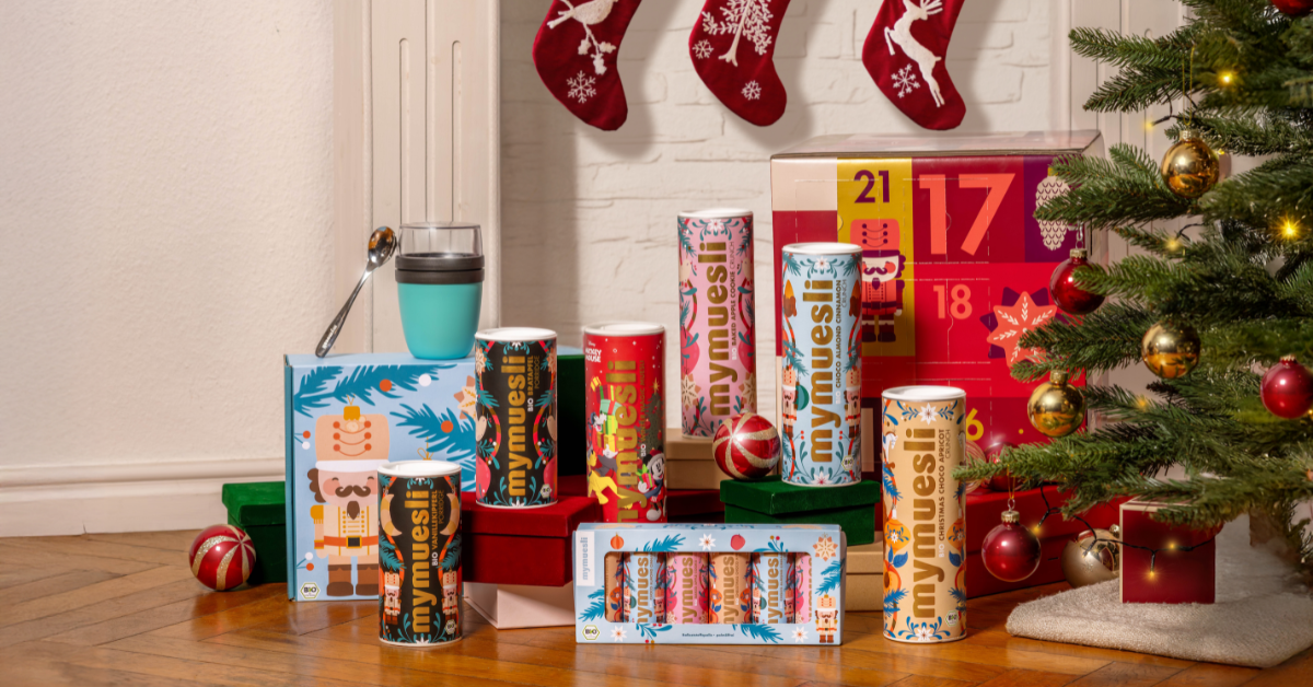 Das mymuesli Weihnachtssortiment, bestehend aus Weihnachtsmüslis, Porridges, Geschenksets sowie der mymuesli Adventskalender. Die Geschenke stehen unter einem geschmückten Weihnachtsbaum, vor einem Kamin. 