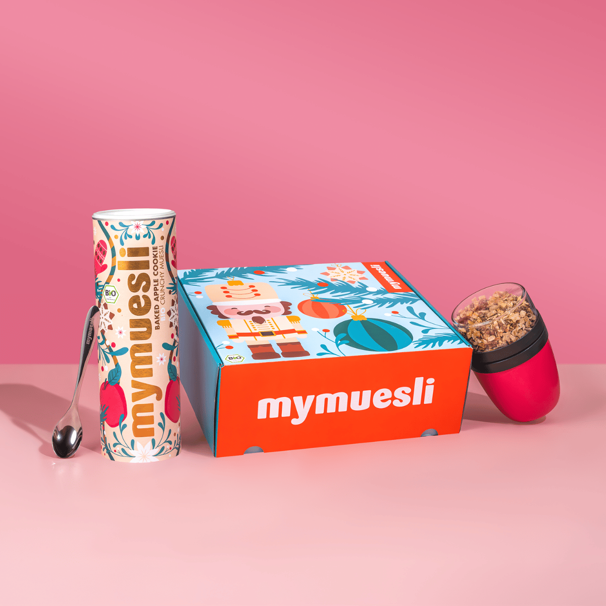 Das mymuesli Geschenkset, bestehend aus Bio Baked Apple Cookie Crunchy, Müsli To-go Becher in magenta / pink und einem silbernen mymuesli Löffel. Verpackt in einer weihnachtlichen Geschenkbox. 