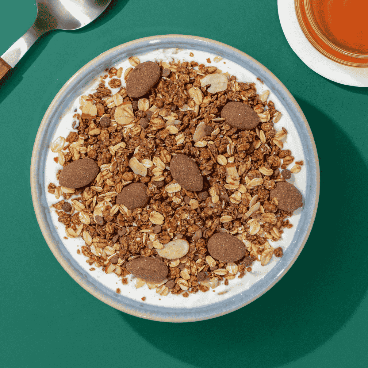 Müslischale mit Schokoladigem Müsli auf einem grünen Hintergrund. Nebendran liegt ein Löffen und eine Tasse Tee.