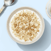 Eine helle Schüssel mit weißem Joghurt und hellem Knuspermüsli, von oben fotografiert auf hellblauem Untergrund.