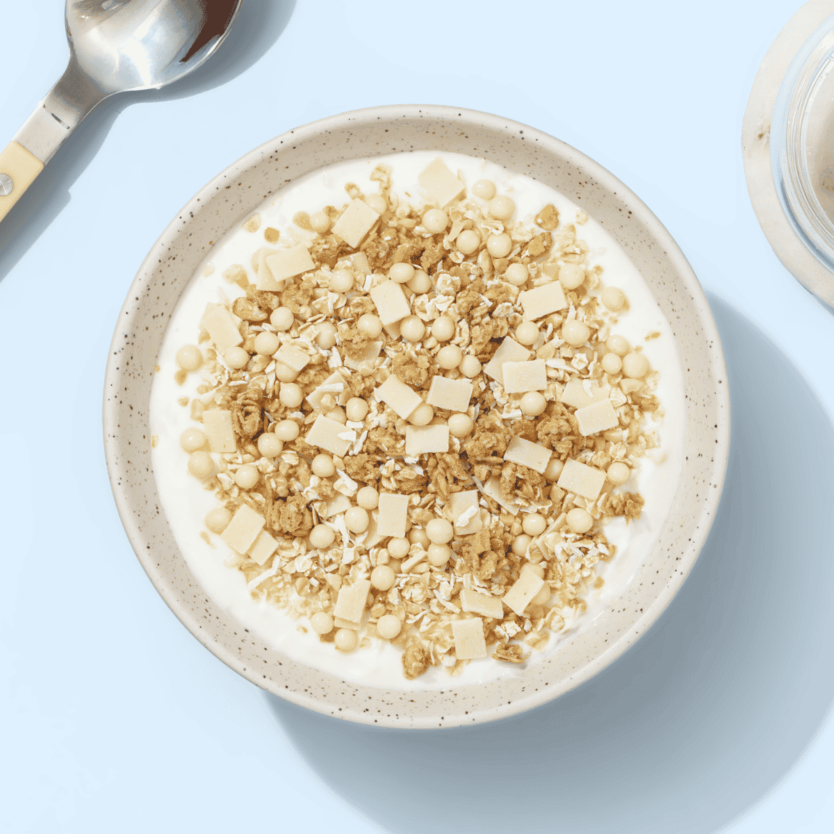 Eine helle Schüssel mit weißem Joghurt und hellem Knuspermüsli, von oben fotografiert auf hellblauem Untergrund.