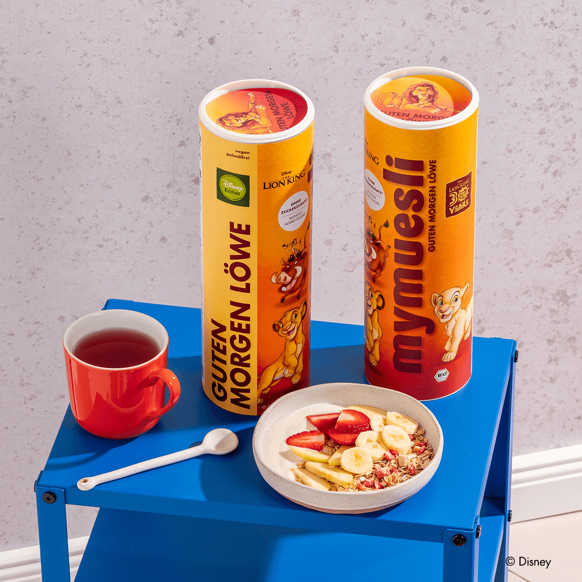 Zwei Disney Guten Morgen Löwe Müsli Dosen, das Müsli in einer Müslischale mit Bananenscheiben, Erdbeeren, Apfelspalten und Joghurt, daneben eine Tasse Tee