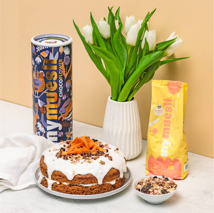 Naked Carrot Cake auf einem Teller, eine Müslidose und Blumen daneben