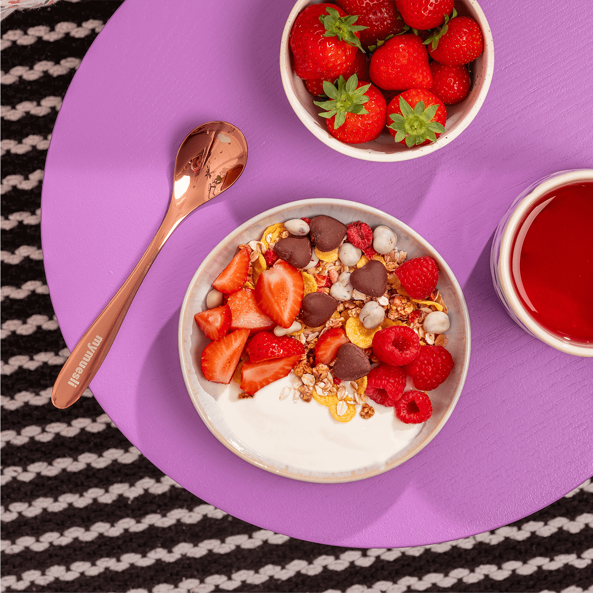 Frühstücks-Ensemble auf lila Untergrund: Eine Joghurt-Bowl mit Erdbeeren und Schokomüsli, dazu ein kupferfarbener Löffel und frische Erdbeeren.