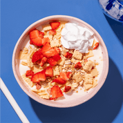Eine rosa Schüssel mit hellem Müsli, Erdbeerstücken und einem Klecks Sahne oder Joghurt auf einem blauen Untergrund.