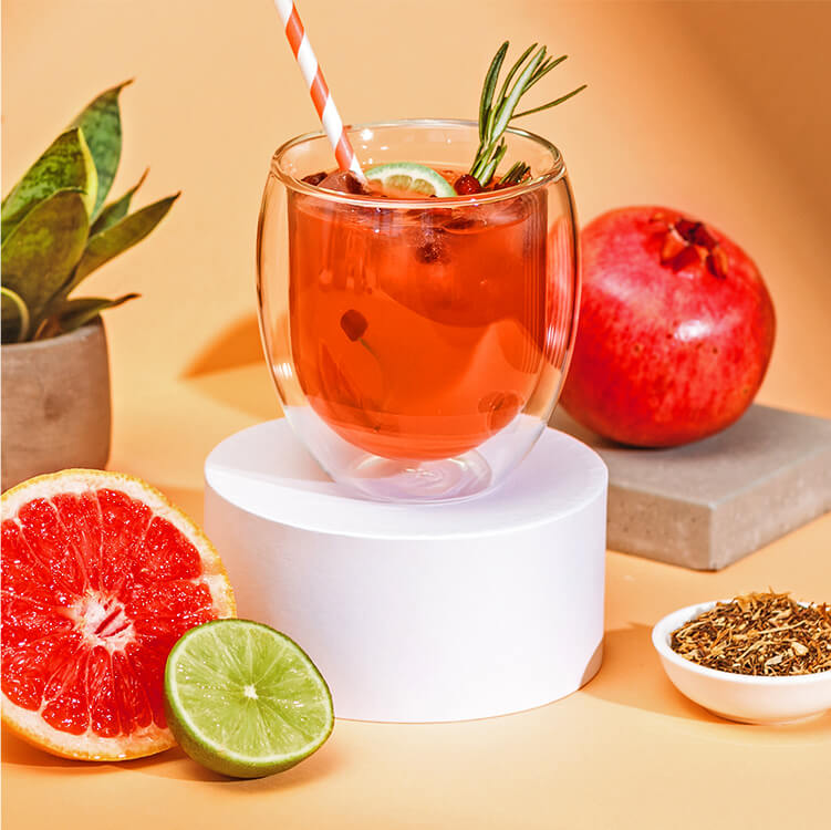 Grapefruit Granatapfel Eistee in einem Glas und daneben eine halbe Grapefruit, Limette, Tee und Granatapfel