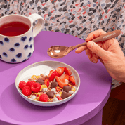 Eine Hand hält einen kupferfarbenen Löffel über eine Schüssel mit Joghurt und Früchten, die auf einem lila Tisch neben einer gepunkteten Tasse steht.