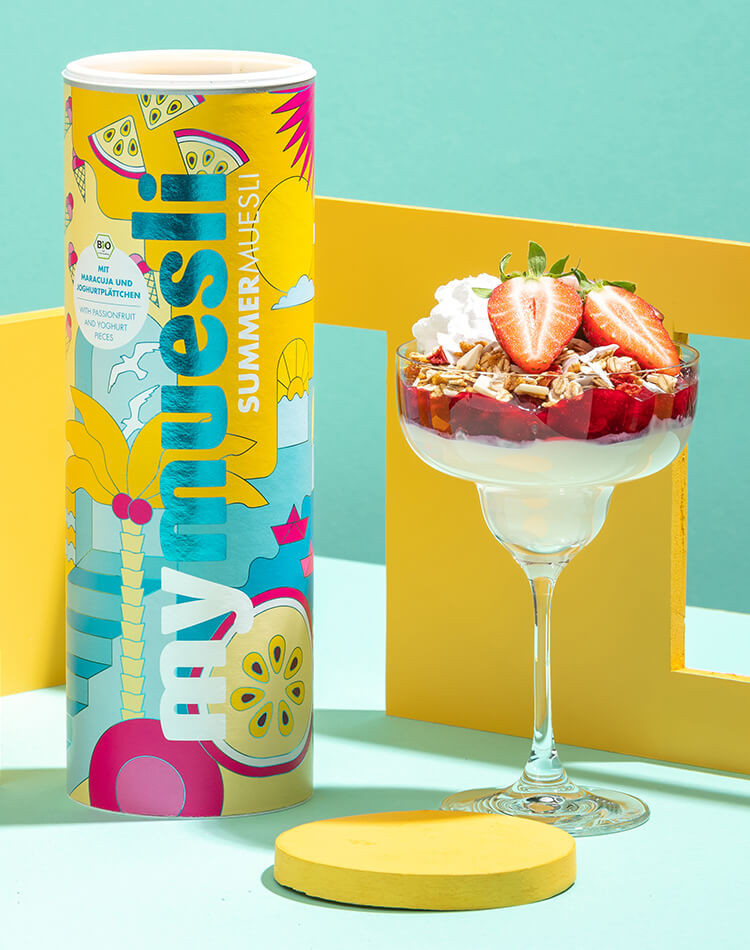 Buntes mymuesli Summer Muesli mit glaenzender Aufschrift neben Cocktailglas mit Joghurt und Muesli